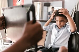 A young boy in a barbershop chair examines his new haircut using a handheld mirror.